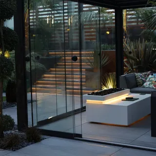 Gardens of Elegance designs and composes luxury outdoor living spaces that blur the line between indoors and out. At the heart of this feature is a bespoke glass-room pergola with sleek sliding glass 